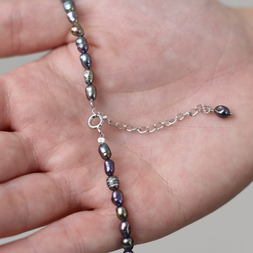 
                      
                        Close-up of a hand holding a choker necklace with black rice-shaped baroque pearls and a rhodium-plated silver chain.
                      
                    