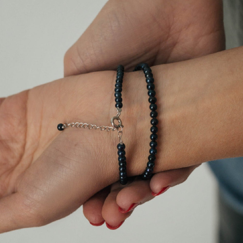 
                      
                        Choker with round black pearl worn as a wristlet on a person's wrist, featuring a minimalist design with a silver chain.
                      
                    